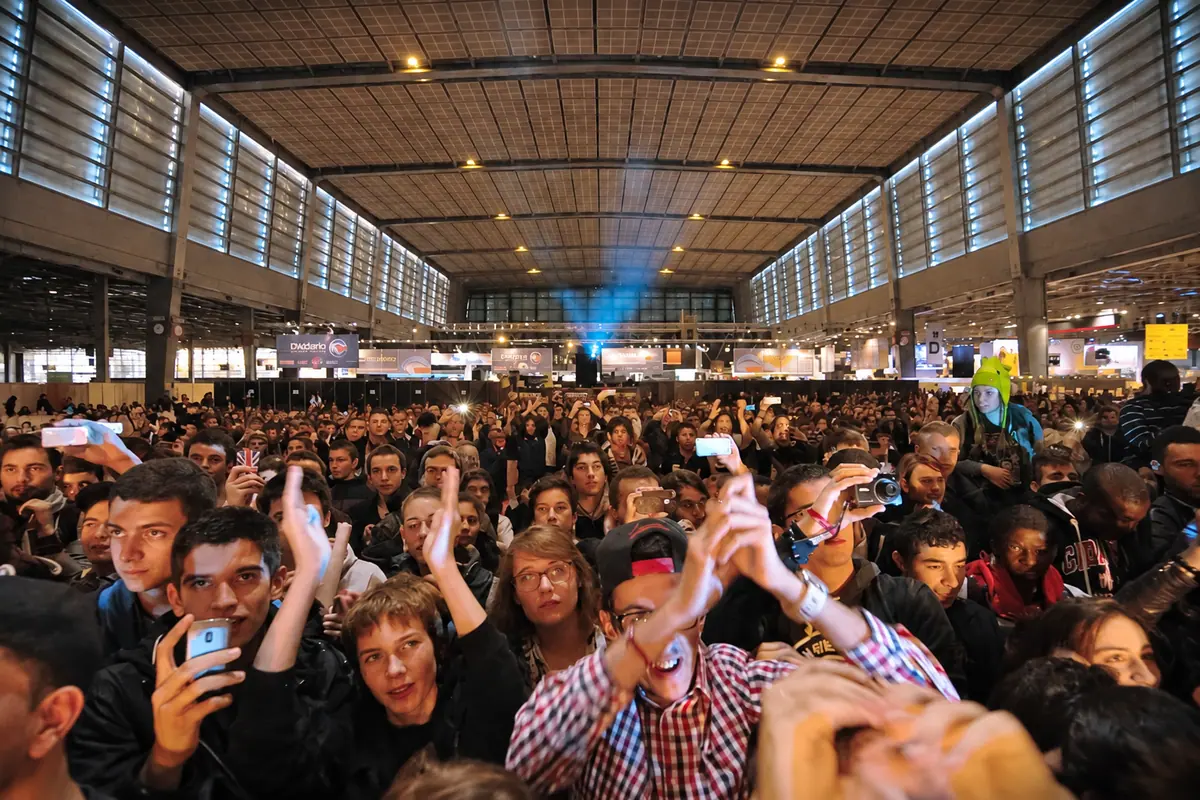 Une foule compacte dans une grande halle, téléphones levés vers la scène.